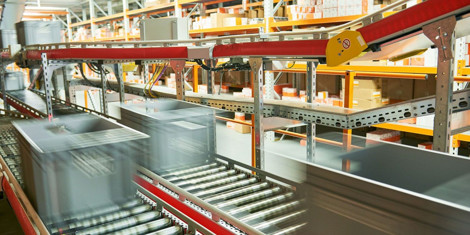 Plastic bins move quickly along a conveyor belt system in a warehouse, with shelves of boxes and packages in the background—showcasing an automated logistics center ideal for advanced automation analysis.