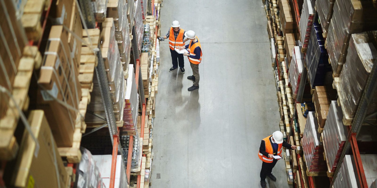 Three workers in orange safety vests and white helmets inspect shelves in a large warehouse filled with stacked boxes and packages, viewed from above.