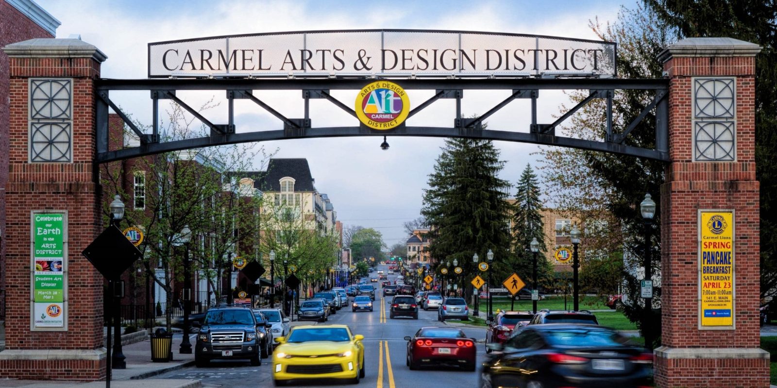 A street view of the Carmel Arts & Design District entrance with a large arched sign overhead, cars driving by, trees, brick pillars, and city buildings visible in the background.
