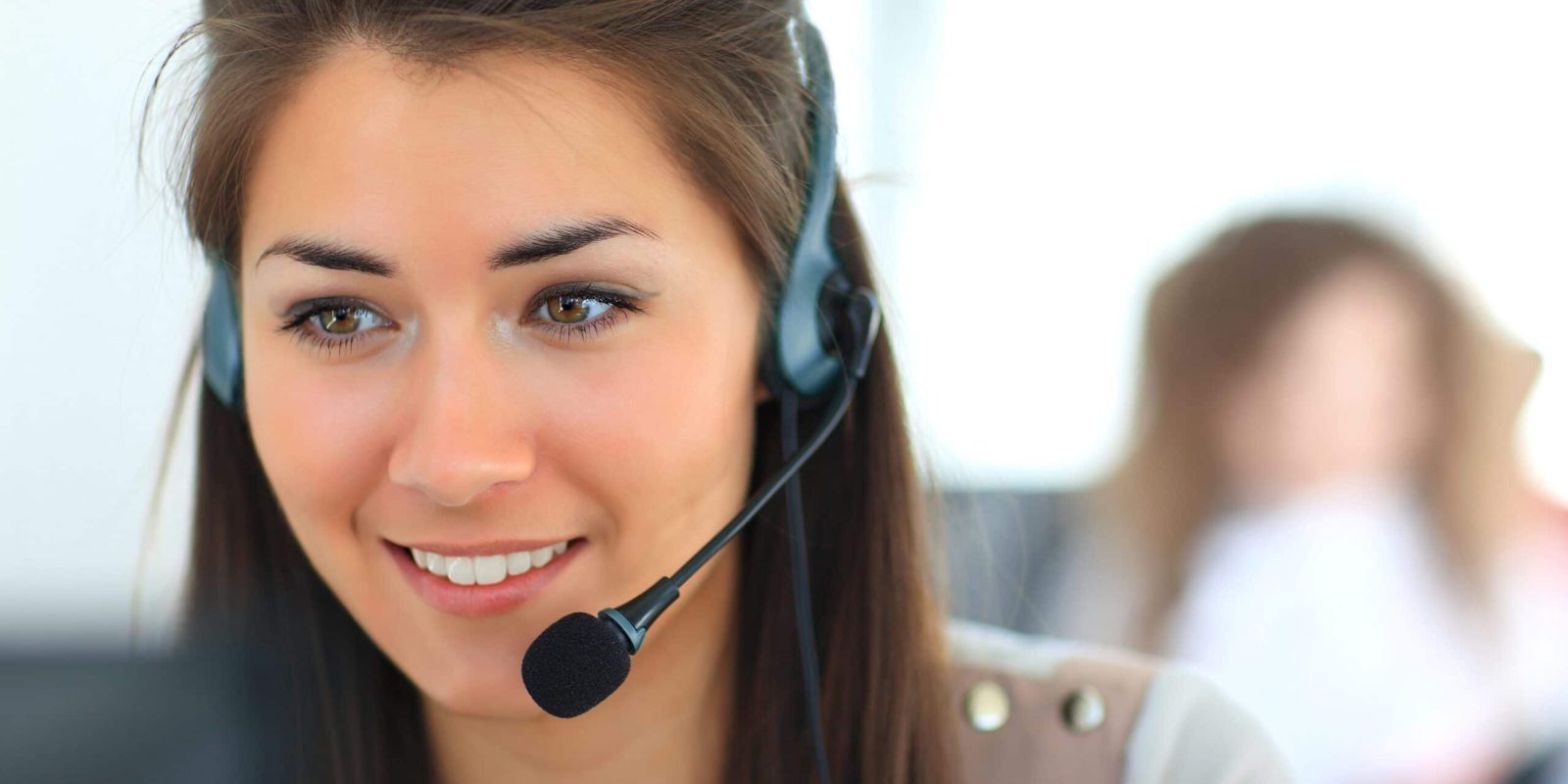 A woman wearing a headset with a microphone is smiling while looking at a computer screen, suggesting she is working in a customer service or call center environment. The background is blurred.