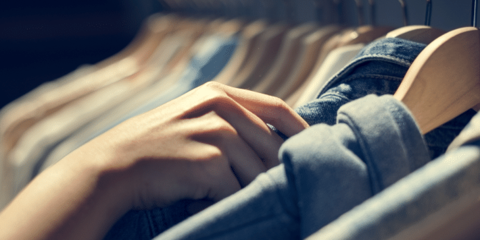 A person’s hand is browsing through a row of clothes on hangers, selecting or inspecting a garment in a softly lit store or closet.