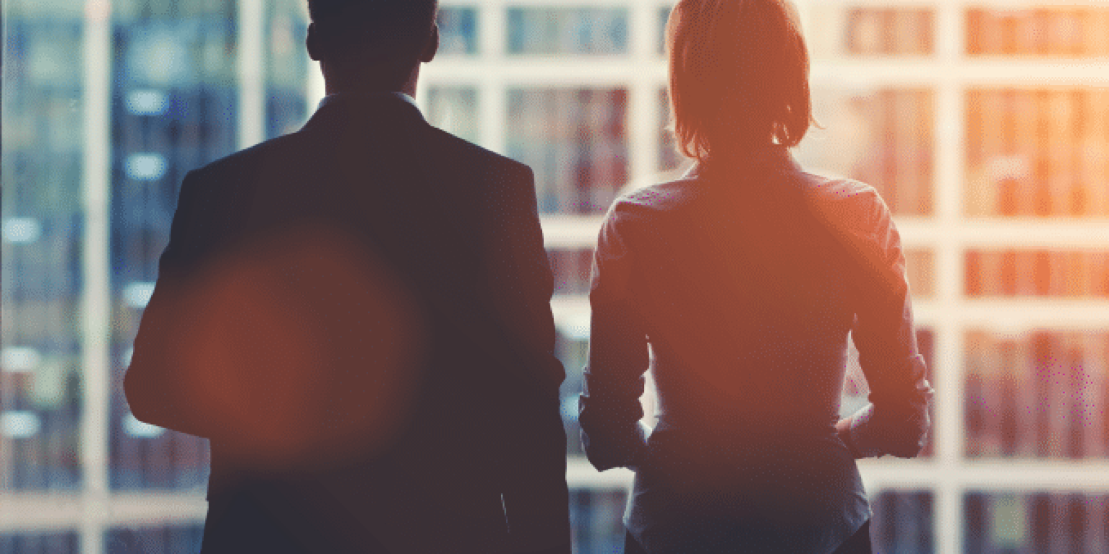 Two business professionals, a man and a woman, are seen from behind standing in front of a large window overlooking tall office buildings in a city, with sunlight streaming in.