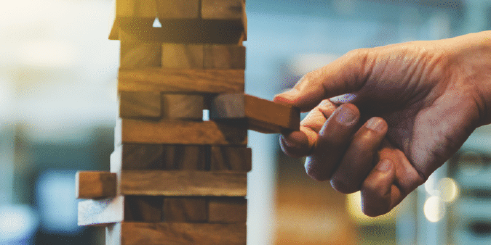 A hand carefully pulls a wooden block from a stacked Jenga tower on a table, with blurred indoor background and warm lighting.