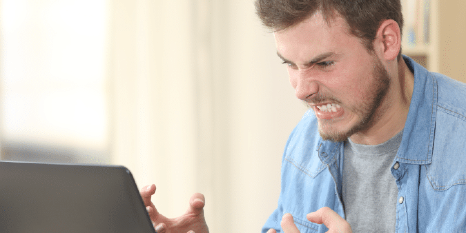 A man sitting at a desk in front of a laptop clenches his fists and grimaces in frustration. Papers, a cup, and a smartphone are on the desk, and bookshelves are in the background.