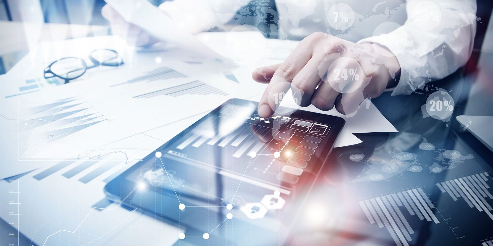 A person reviews financial charts on paper and a digital tablet at a desk, surrounded by graphs and analytics overlays, representing business analysis or data-driven decision-making with advanced technology solutions.