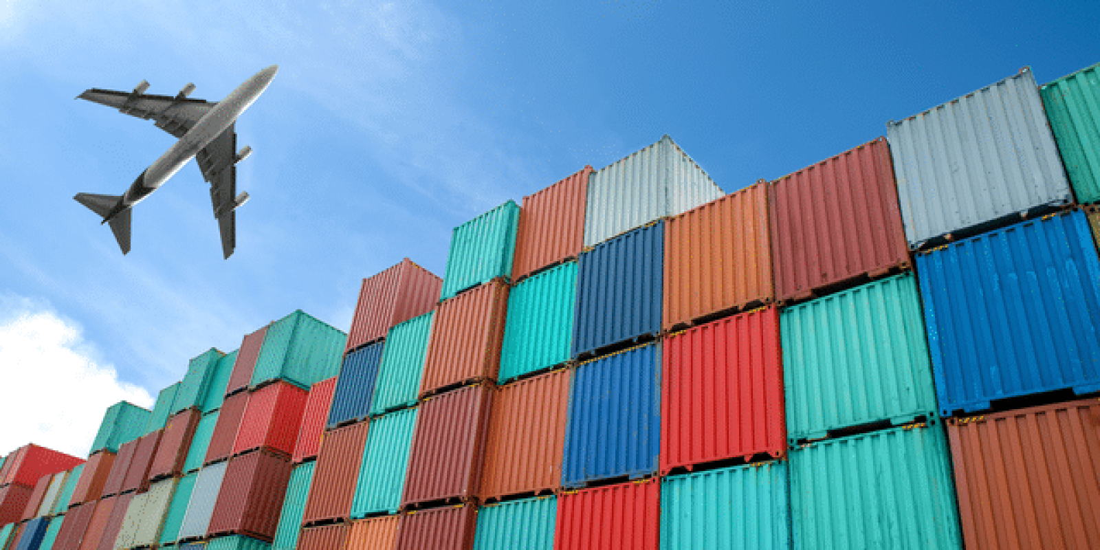 A commercial airplane flies above a large stack of colorful shipping containers under a clear blue sky.