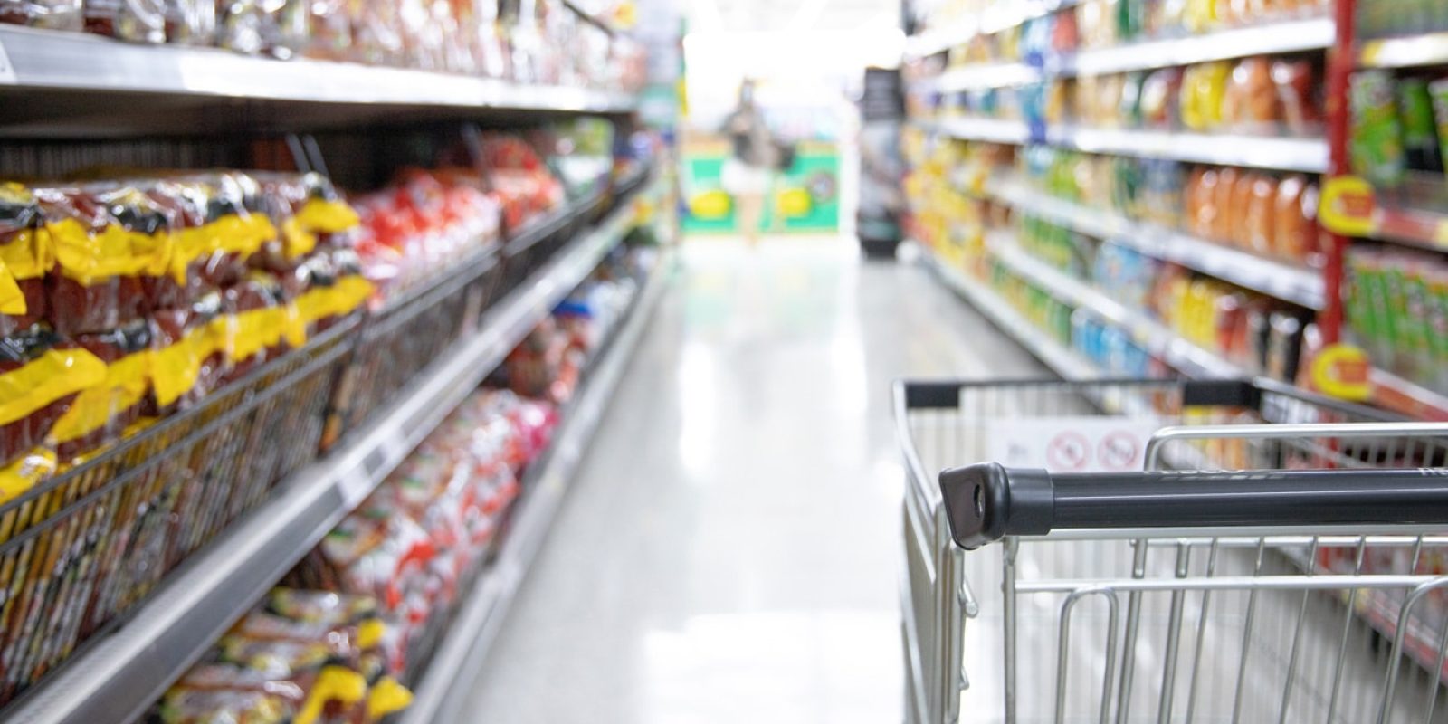A shopping cart in a grocery store aisle, surrounded by shelves stocked with various packaged food items on both sides. The scene is brightly lit, and the shelves appear well-organized.