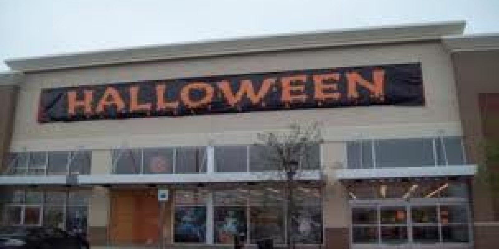 A large store with a black banner above the entrance that reads HALLOWEEN in orange, festive letters. The storefront has glass windows and doors, with some Halloween decorations visible inside.