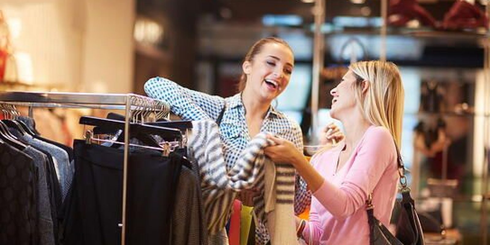 Two women smiling and laughing while shopping for clothes in a brightly lit store, holding and looking at a striped sweater near a rack of clothing.