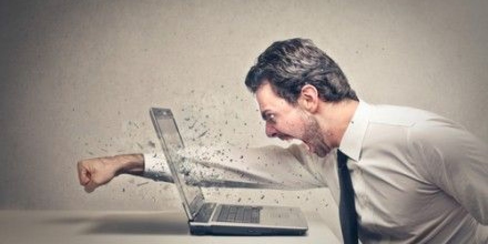 A man in a dress shirt and tie angrily punches a laptop, causing the screen to shatter and fragments to fly, while sitting at a desk against a plain background.