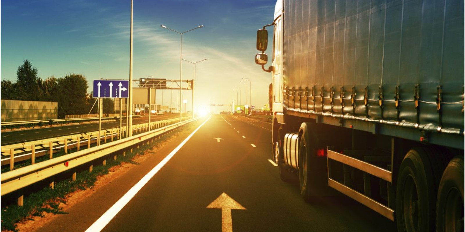 A large truck drives down a highway at sunrise or sunset, with bright sunlight ahead, road signs on the left, and a clear sky overhead. The road features lane markings and guardrails.
