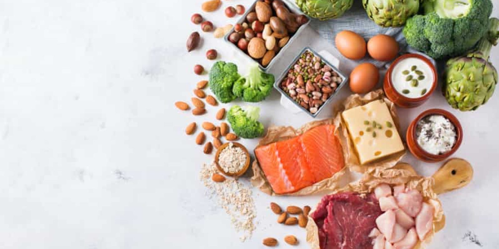 Assorted healthy foods including raw salmon, beef, chicken, cheese, eggs, nuts, seeds, broccoli, artichokes, yogurt, and legumes arranged on a light surface with space on the left.