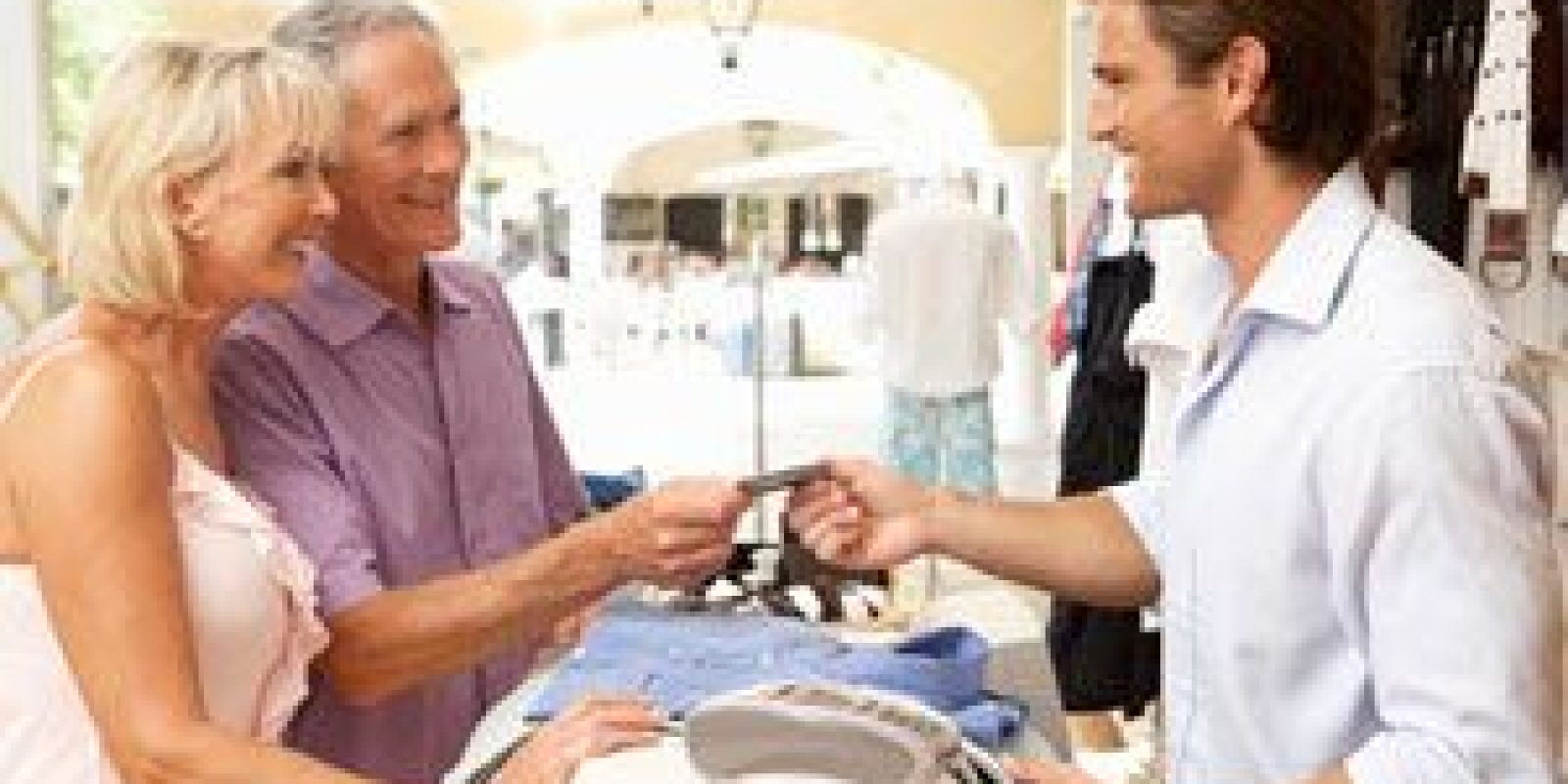 An older couple stands at a store counter, smiling as the man hands a credit card to a young male cashier. Clothes and a credit card terminal are visible on the counter in a bright, open shop.