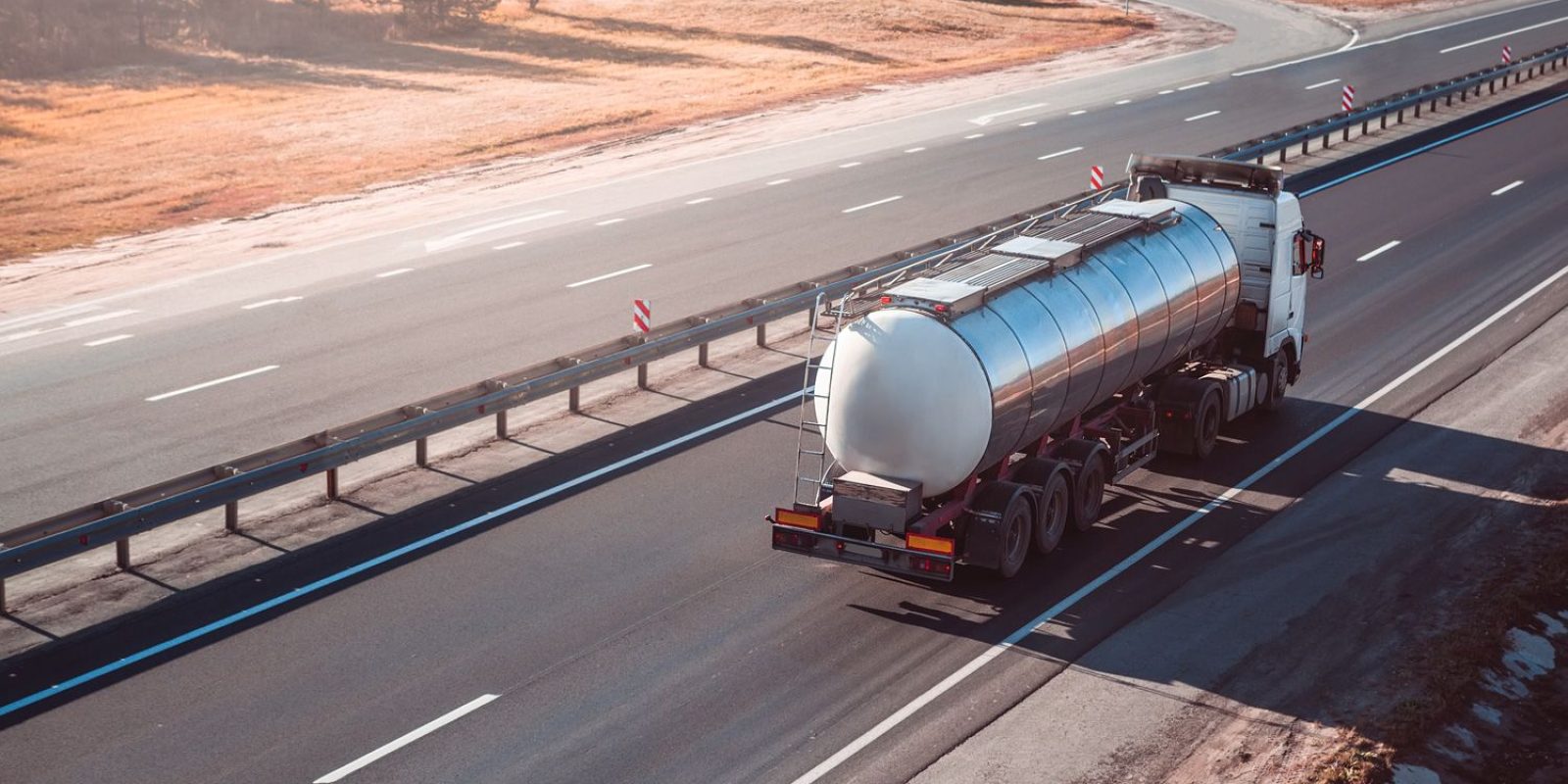 A white tanker truck drives on a mostly empty multi-lane highway bordered by grass and guardrails, with clear weather and gentle sunlight.