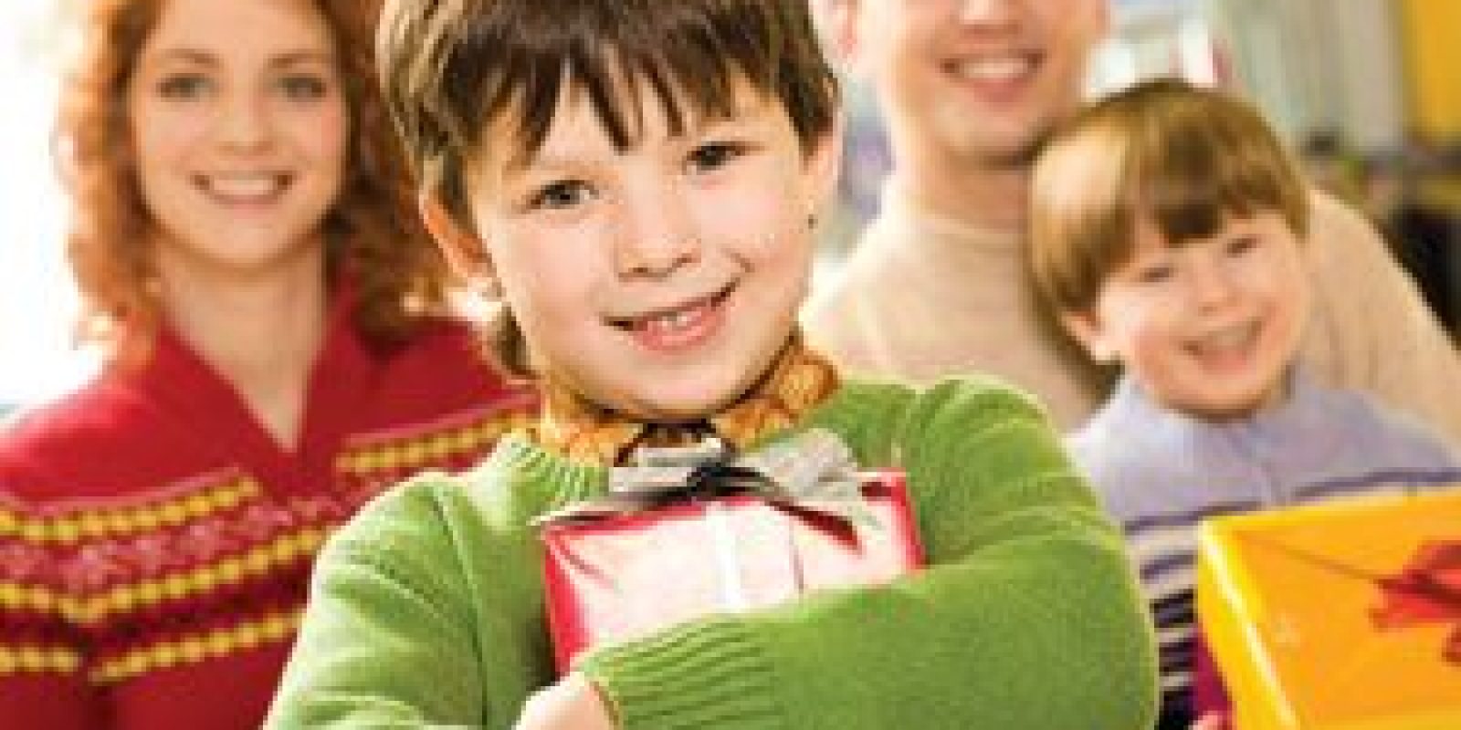 A smiling young boy in a green sweater holds a wrapped gift, standing in front of a smiling woman, man, and another young boy who is also holding a present. The group appears happy and festive.