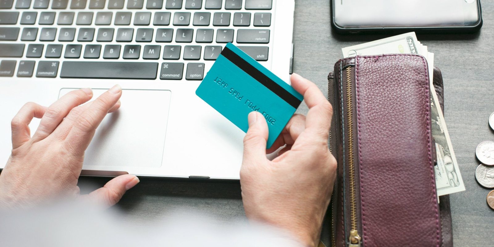 A person holding a blue credit card sits at a laptop, with a purple wallet, cash, coins, and a smartphone on the table nearby. The scene suggests online shopping or banking.