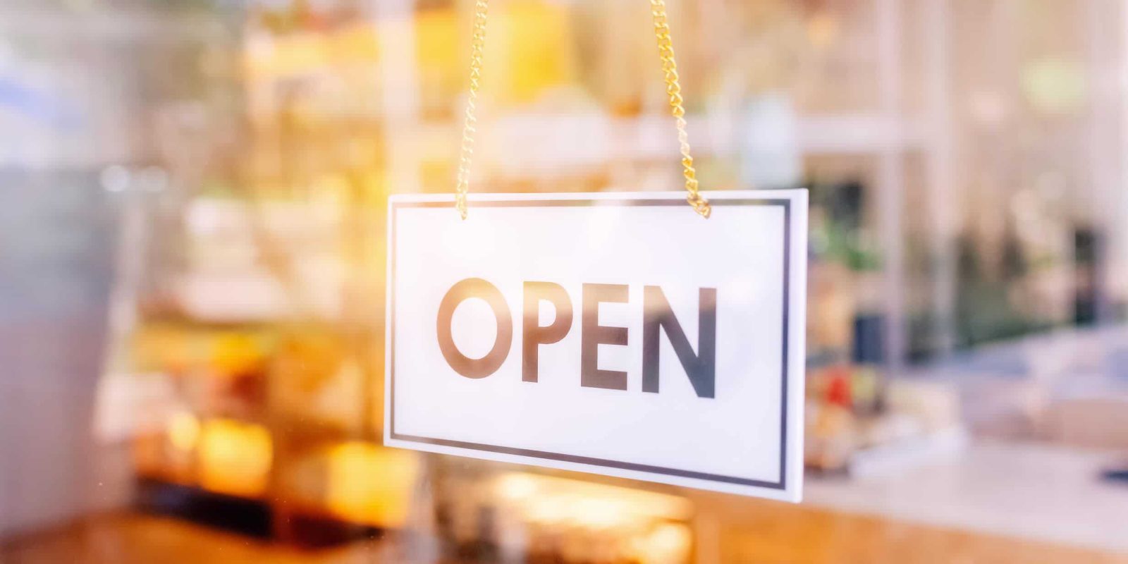 A white rectangular sign with the word “OPEN” in bold black letters hangs on a glass door, with a blurred interior of a shop or café visible in the background.