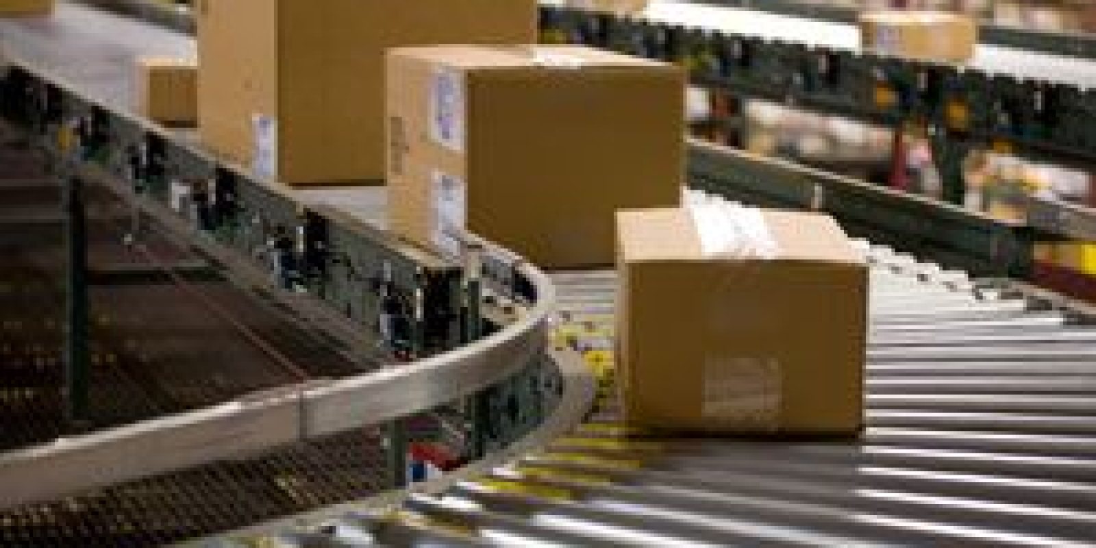 Several cardboard boxes move along a curved conveyor belt in a warehouse or distribution center, with shelves and additional packages visible in the background.
