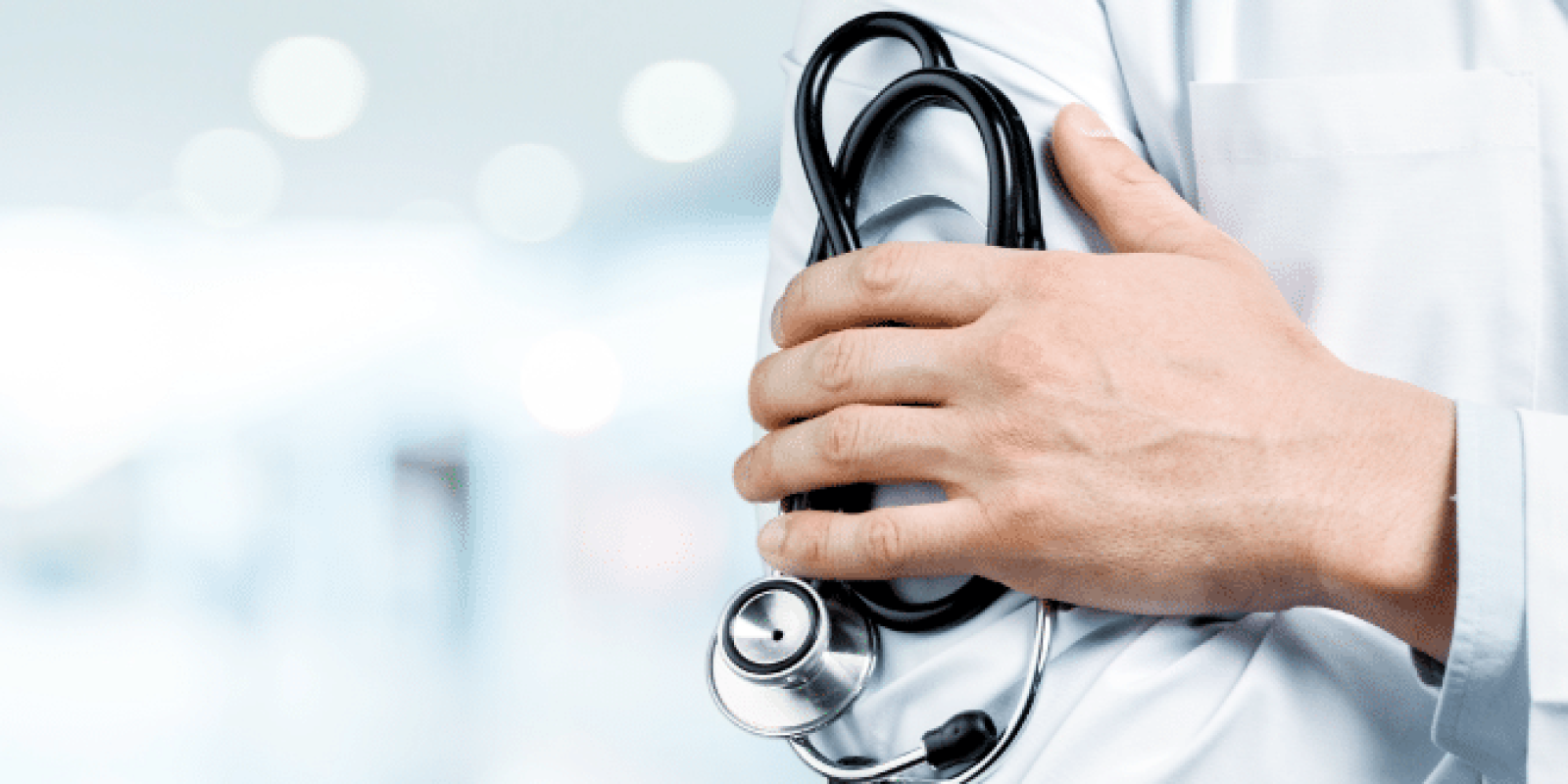 A doctor in a white coat stands with arms crossed, holding a stethoscope, in a bright, blurred medical or hospital setting.