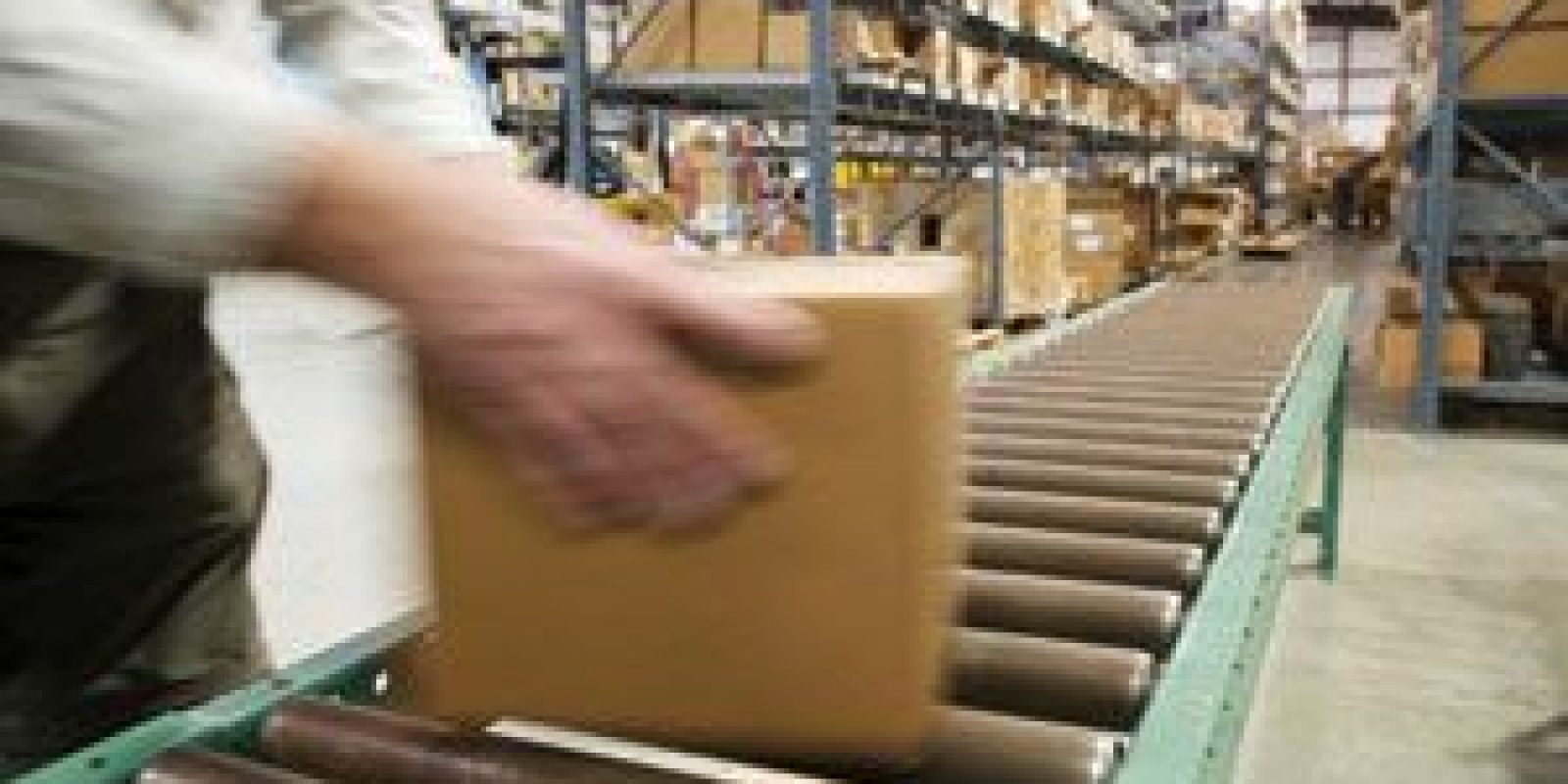 A person places a cardboard box on a conveyor belt in a warehouse filled with shelves of packed boxes.