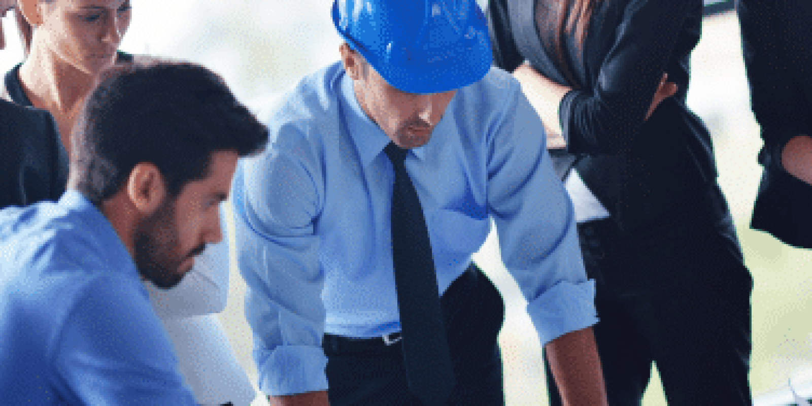 Five professionals, including one person in a blue safety helmet, closely examine blueprints or plans on a table in a bright office, appearing focused and engaged in discussion.