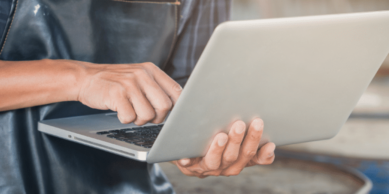 A person wearing a dark apron and plaid shirt uses a laptop, standing in what appears to be a workshop or industrial setting. Only the torso and hands are visible.