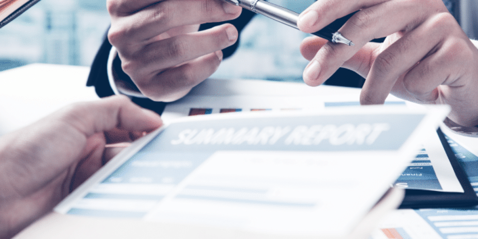 Two people in business attire review a printed summary report at a desk. One person gestures with a pen while the other holds the document, suggesting a discussion or meeting in a professional office setting.