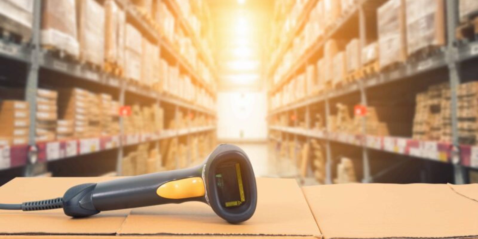 A barcode scanner rests on stacked cardboard boxes in a warehouse with shelves full of inventory management supplies, brightly lit by sunlight in the background.
