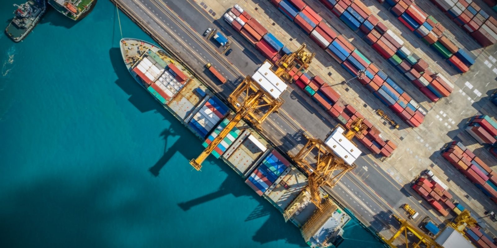 Aerial view of a shipping port with large cargo ships docked, yellow cranes loading containers, and stacks of colorful shipping containers—highlighting the vital role ports play in global supply chains—lined up along the dock next to bright blue water.