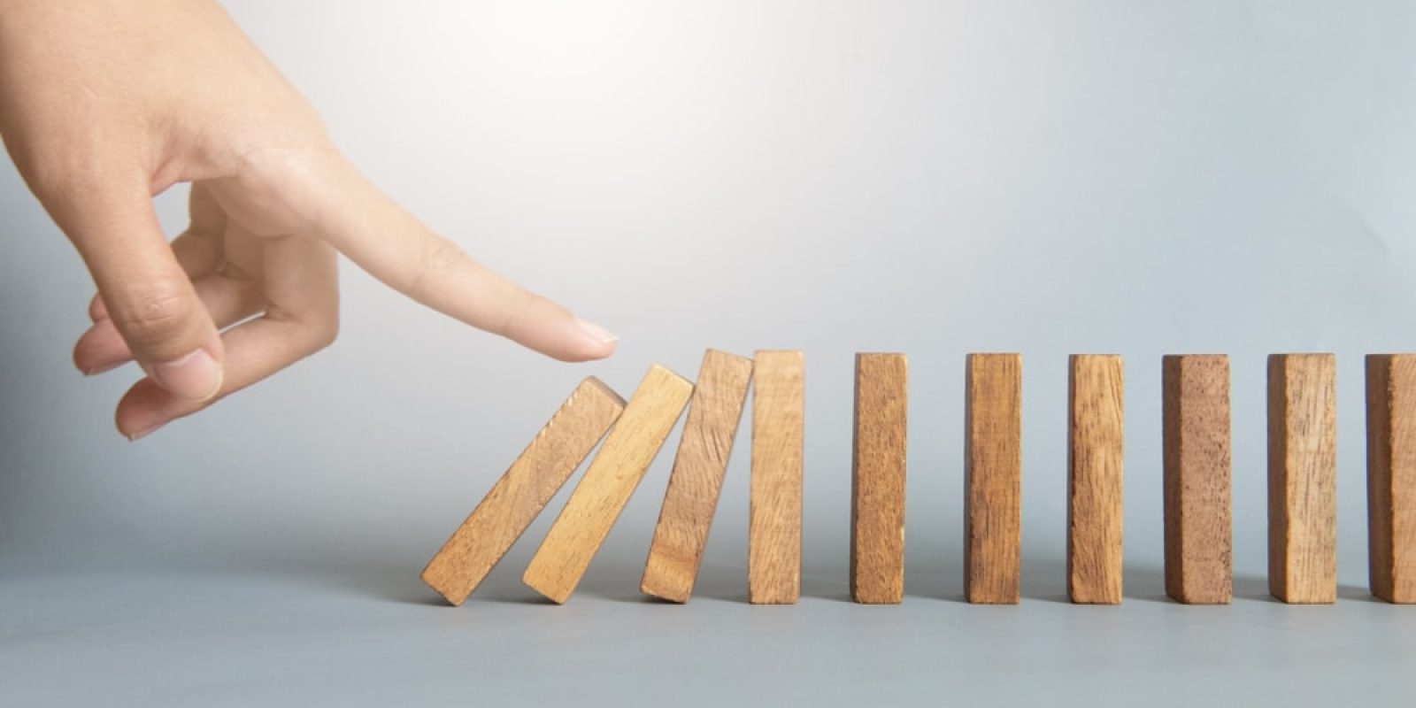 A hand is about to push the first of several standing wooden dominoes, illustrating a bullwhip effect chain reaction. The dominoes are arranged in a line on a smooth surface against a plain background.