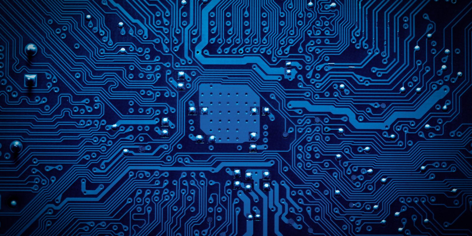 Close-up view of a blue printed circuit board showing intricate pathways and connections, with small metallic components and a central chip area, representing electronic technology.