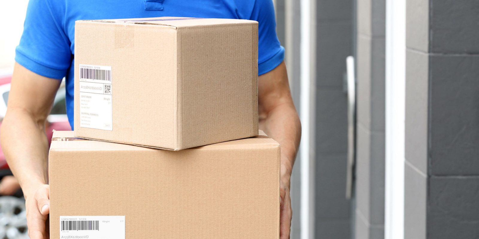 A person in a blue shirt holds two cardboard delivery boxes, standing outside a building with gray walls and a door. Both boxes have shipping labels attached.