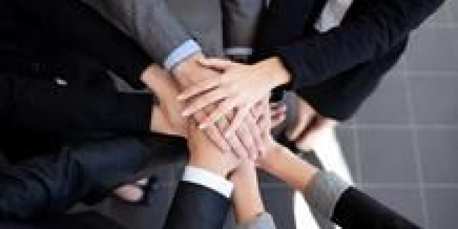 A group of people wearing business attire stand in a circle and stack their hands together in the center, symbolizing teamwork and unity.