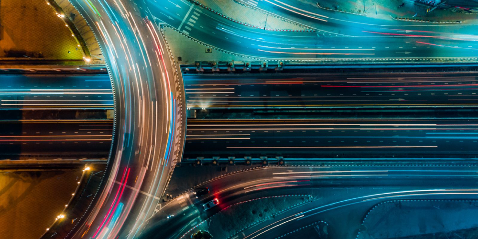 Aerial view of a brightly lit highway interchange at night, with streaks of red and blue lights from moving vehicles creating dynamic, colorful lines across multiple lanes and overpasses.