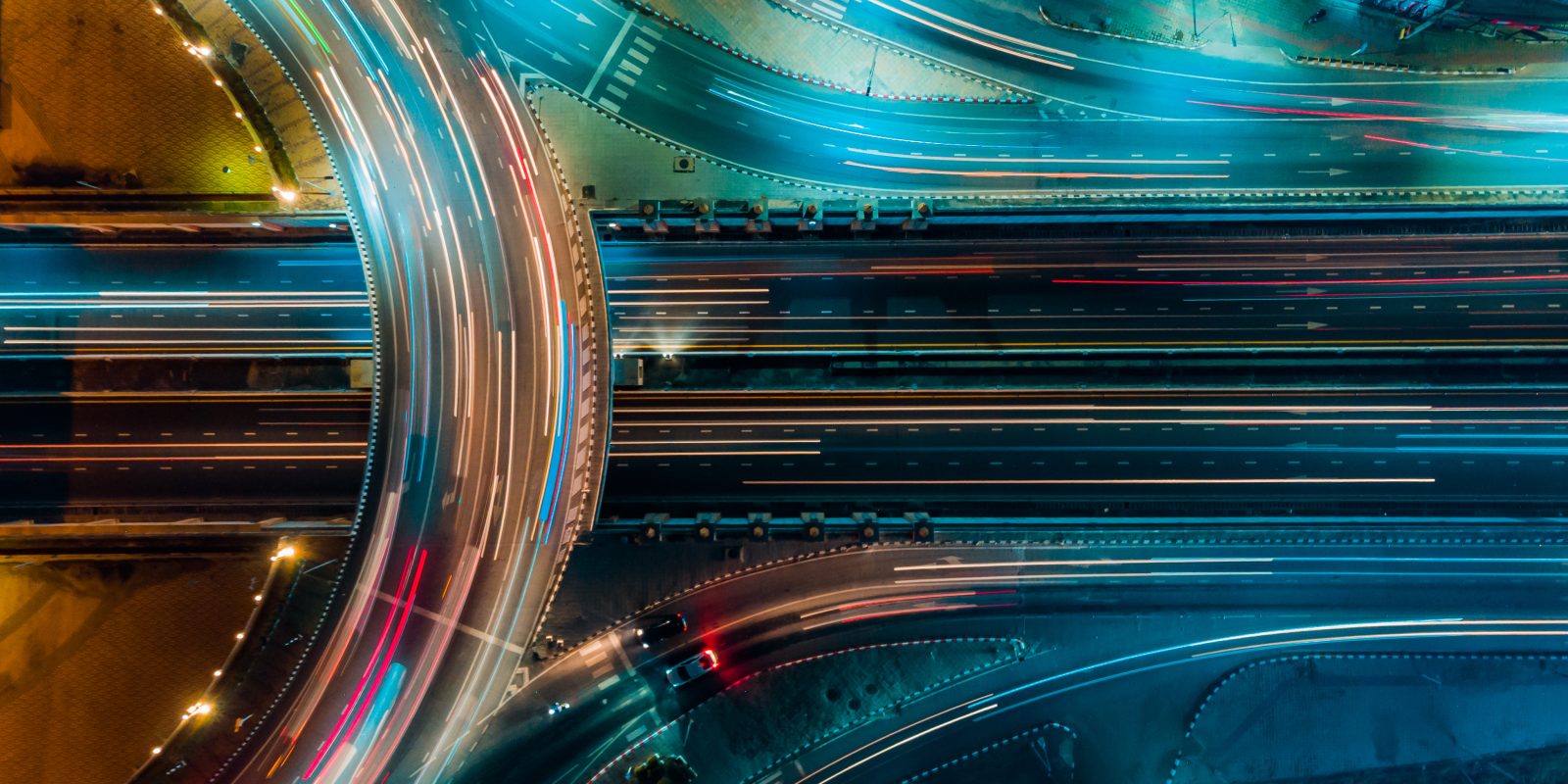 Aerial view of a multi-lane highway interchange at night, with bright blue and yellow streetlights illuminating the roads and blurred light trails from moving vehicles.
