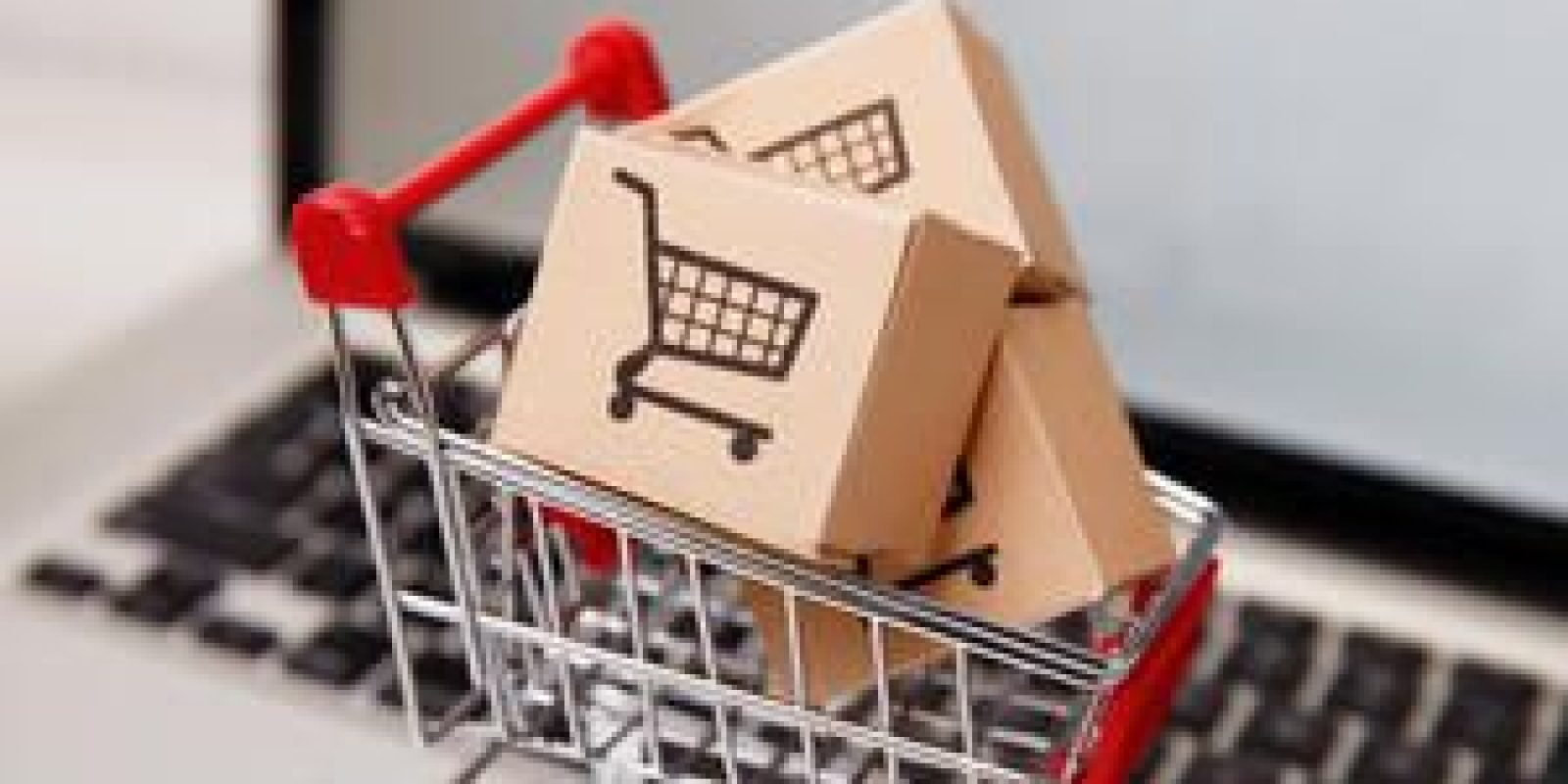A small shopping cart filled with cardboard boxes, each featuring a shopping cart icon, sits on a laptop keyboard, representing online shopping or e-commerce.