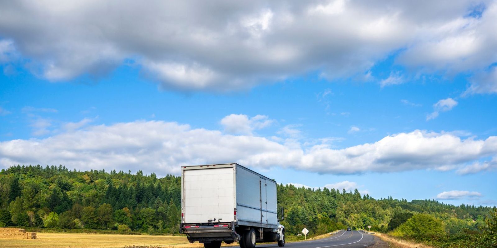 A white semi-truck drives down a curving rural road bordered by green trees and fields under a partly cloudy blue sky.