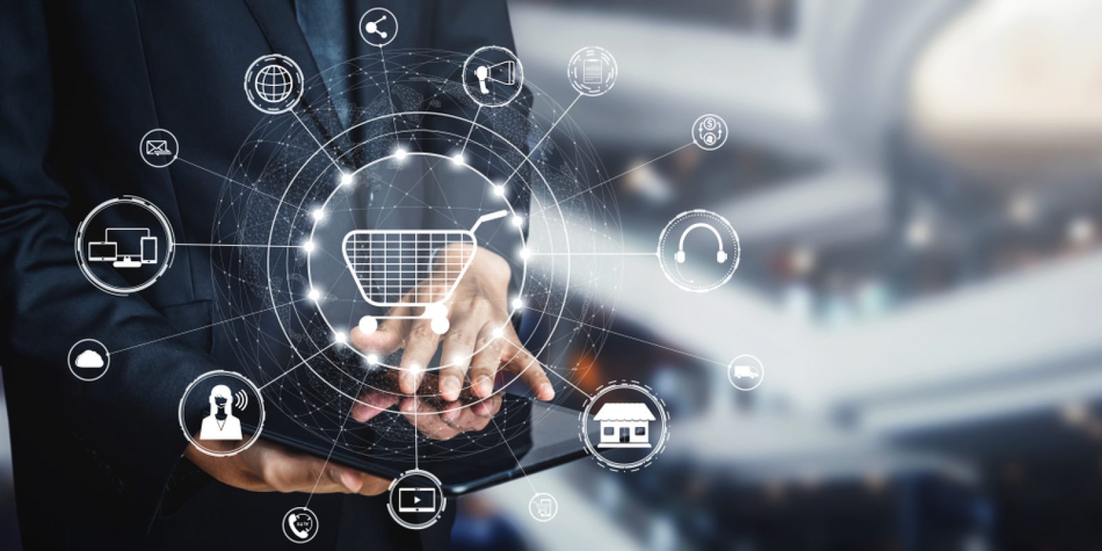 A person in a suit uses a tablet, with a digital overlay of a shopping cart icon surrounded by various e-commerce and technology icons, representing online shopping and digital business concepts.
