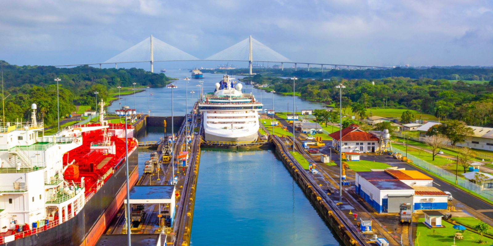 Several large ships pass through a canal lock surrounded by green landscape and buildings focused on warehouse safety, with a long cable-stayed bridge in the background under a partly cloudy sky. Roads and structures line both sides of the canal.