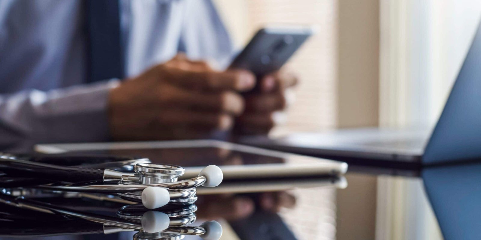 A stethoscope rests on a desk near a tablet and laptop, while a person in business attire uses a smartphone in the background.