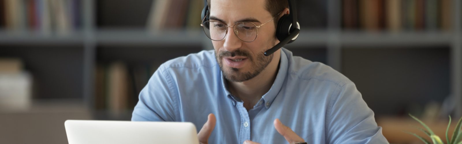 A man wearing glasses and a headset sits at a desk, gesturing as he discusses Microsoft’s Sales Copilot during a video call on his laptop. A bookshelf is visible in the background.