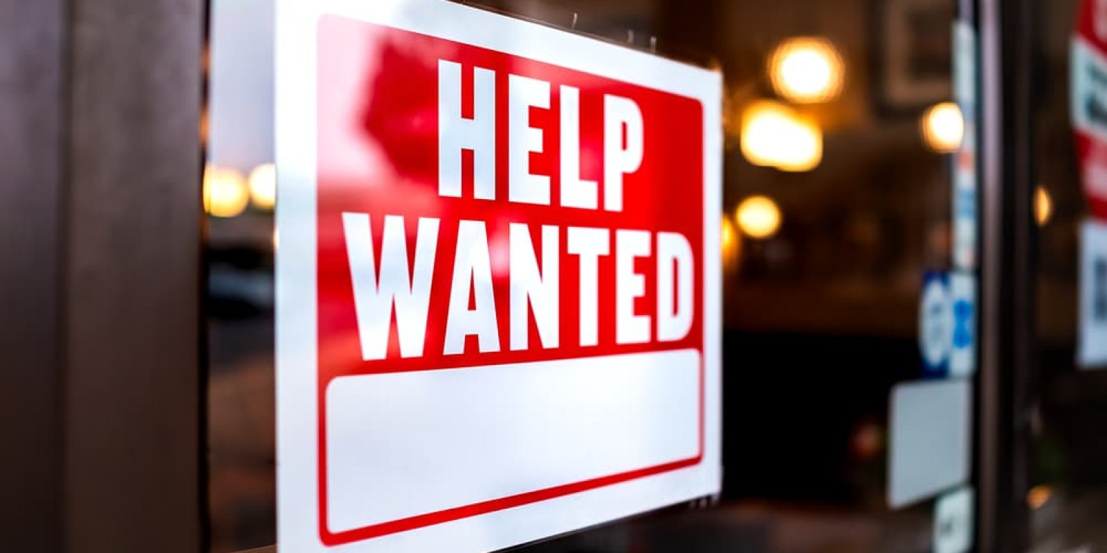 A red and white HELP WANTED sign is displayed in a window, indicating that the business is hiring. The background shows a warmly lit interior with blurred lights.