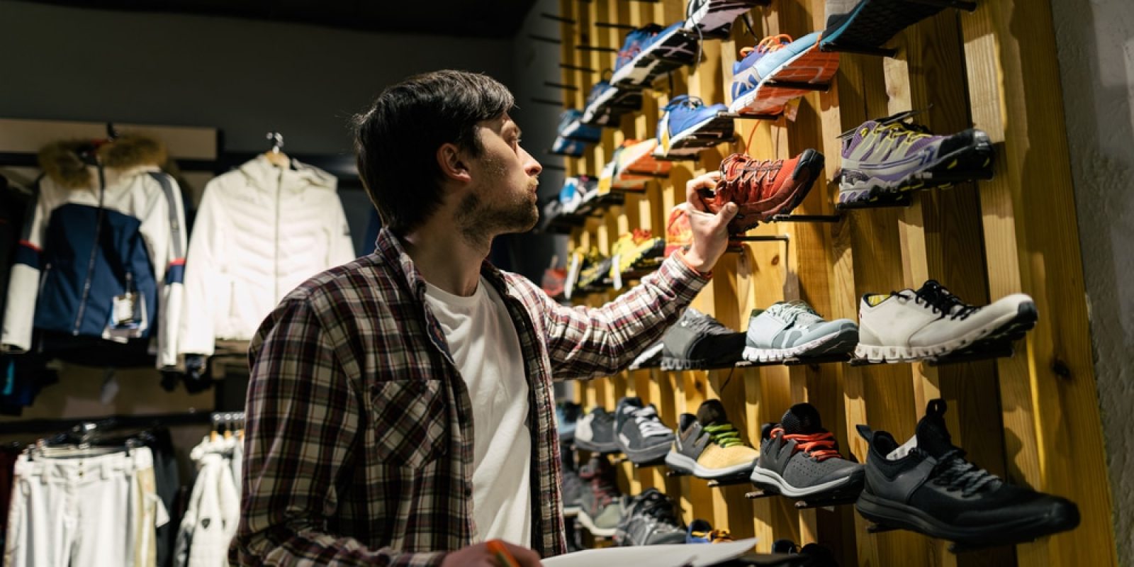 A man in a plaid shirt examines a wall display of athletic shoes in a store, holding a notepad and pen for assortment planning, surrounded by various colorful sneakers and jackets.