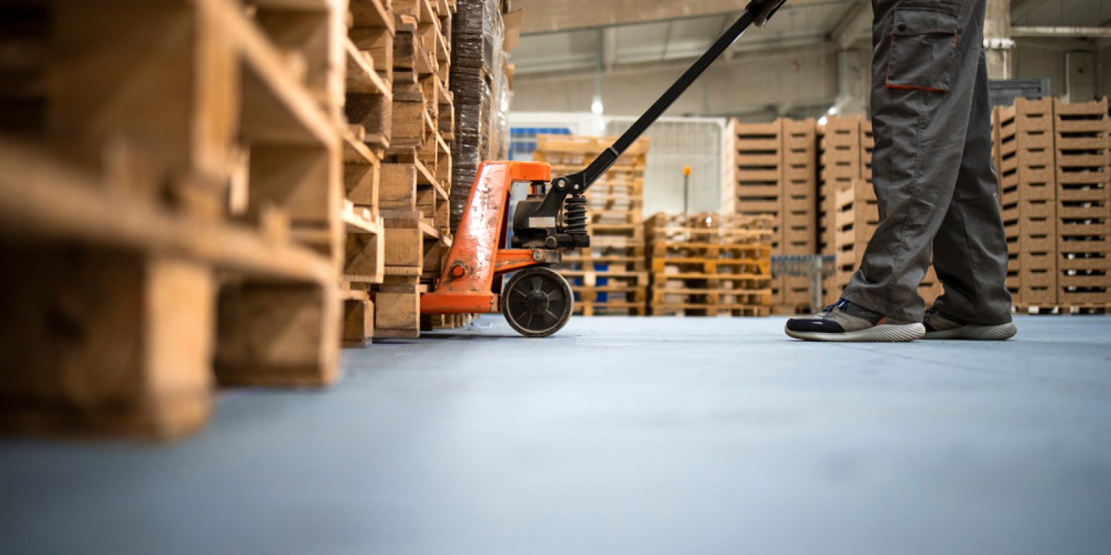 A worker in gray pants and shoes uses a hand pallet jack to move wooden pallets inside a warehouse with stacks of pallets in the background.