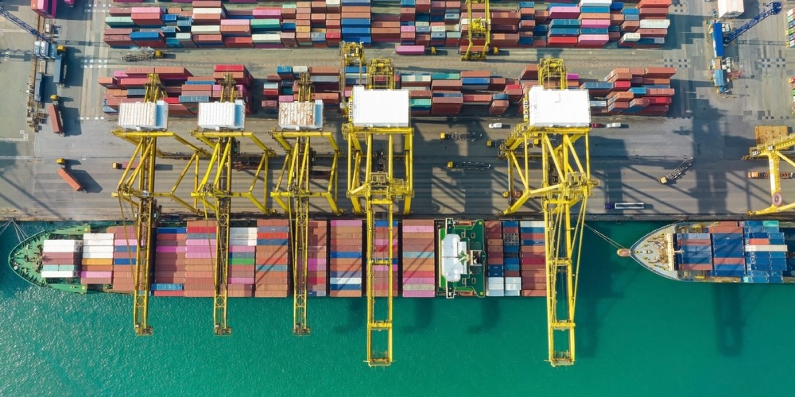 Aerial view of large yellow cranes loading and unloading colorful shipping containers onto cargo ships docked at a busy port, with turquoise water alongside the pier.