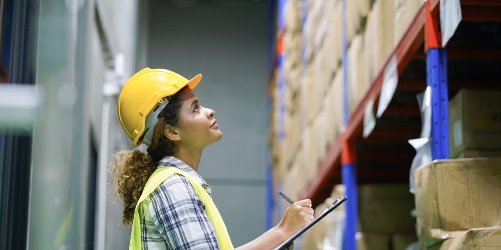 A woman wearing a yellow safety helmet and vest holds a clipboard and pen, looking up at shelves filled with boxes in a warehouse, demonstrating her commitment to warehouse safety.