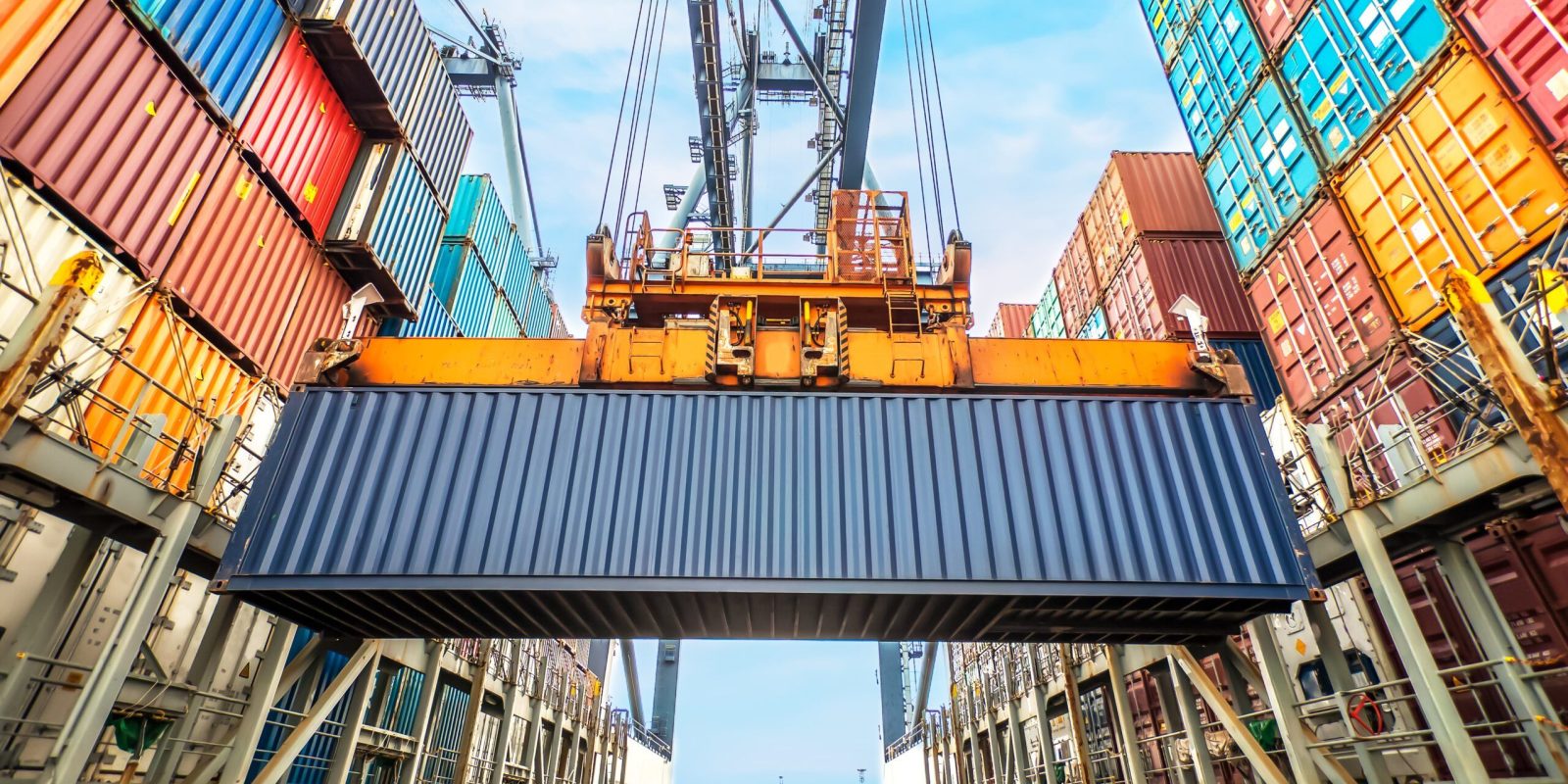 A large shipping container is being lifted by a crane in a busy port, surrounded by stacks of colorful cargo containers against a blue sky.