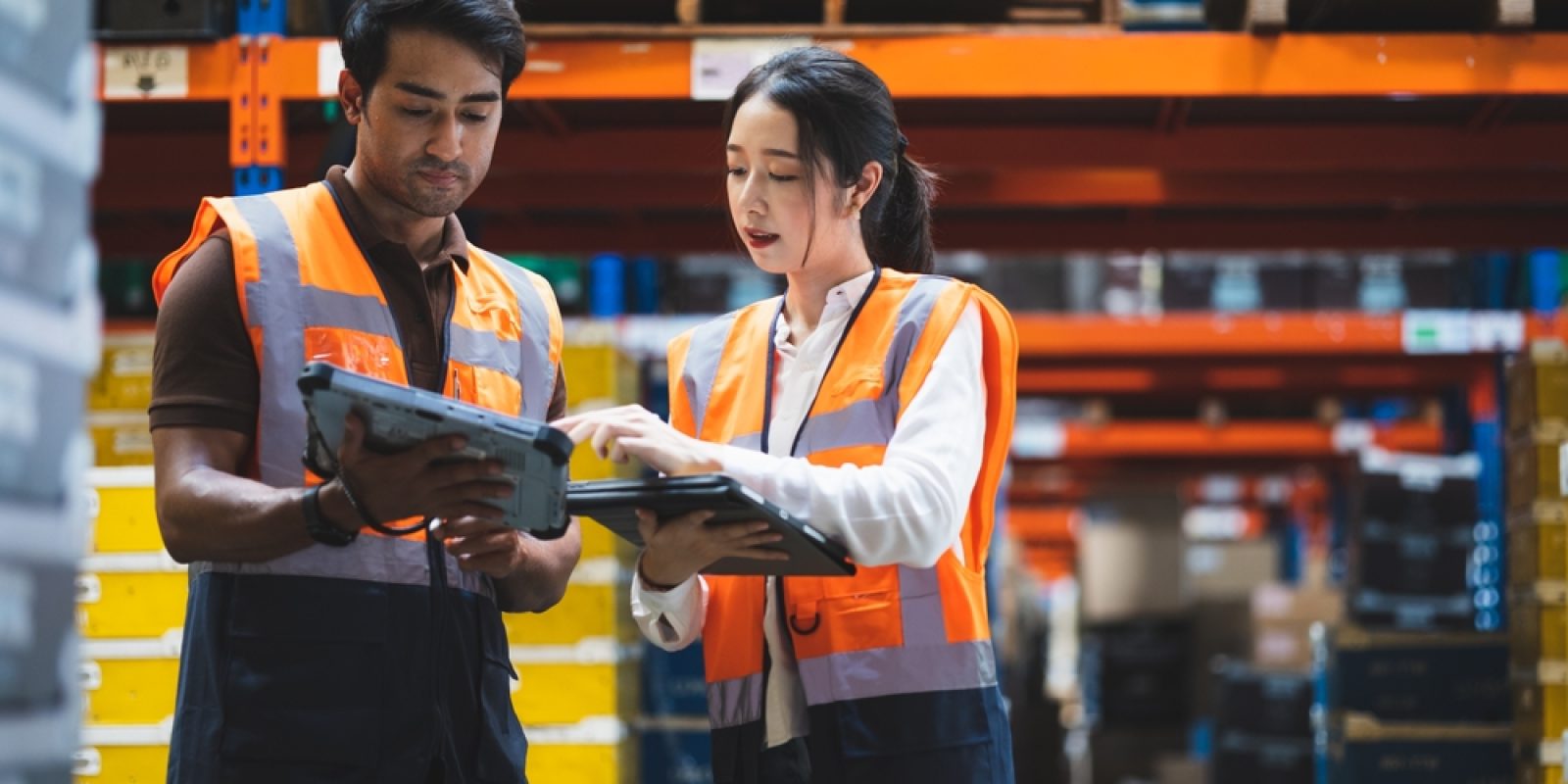 Two warehouse workers wearing orange safety vests review labor management details on a digital tablet, standing among shelves filled with boxes and containers in a storage facility.
