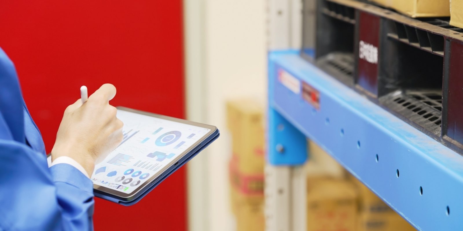 A person in a blue uniform holds a tablet displaying graphs and charts, using a WMS to conduct inventory or inspection in a warehouse filled with shelves and cardboard boxes.