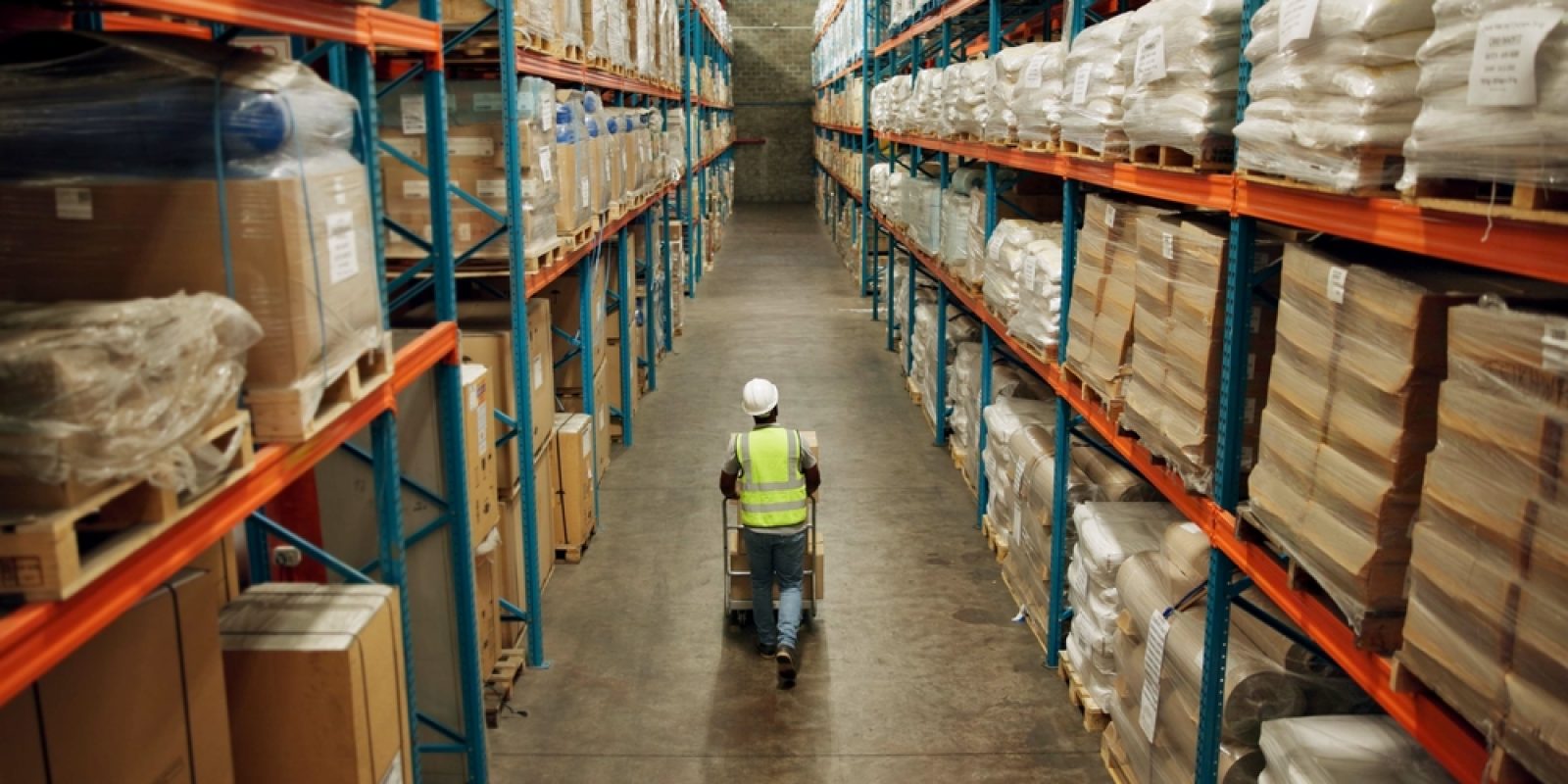 A warehouse worker wearing a reflective vest and hard hat pushes a cart down an aisle lined with tall shelves filled with boxes and wrapped pallets.